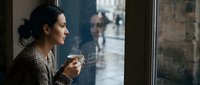 Young woman of South Asian descent with dark hair in a bun, wearing a brown cardigan, holding a warm beverage while gazing thoughtfully out a window on a rainy day, conveying contemplation and solitude.