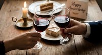 Romantic couple toasting with red wine glasses during intimate dinner celebration with candles, cake and gift on rustic wooden table for anniversary.
