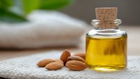 Small glass bottle with cork stopper filled with golden almond oil surrounded by raw almonds on textured fabric for natural skincare and aromatherapy.