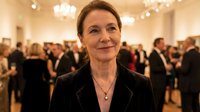 Middle-aged Caucasian businesswoman smiling confidently at corporate event or gallery opening with elegant crowd in background for professional networking content.