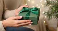 Woman hands holding green gift box with emerald ribbon bow near Christmas tree with warm lights creating festive holiday atmosphere for celebration.