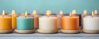 Colorful aromatic candles in glass jars arranged in row on wooden surface, burning with warm glow. Pastel orange, mint green and white create calming spa atmosphere.