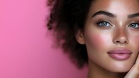 Young African American woman with natural curly hair, freckles and glowing makeup against pink background. Close up beauty portrait showing radiant skin.