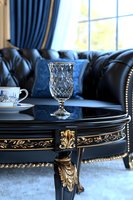 Elegant crystal glass on ornate black and gold coffee table with tufted leather sofa and blue curtains in luxurious classic interior setting.