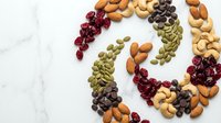 Colorful assorted nuts and dried fruits arranged in decorative spiral pattern on white marble background for healthy snacking and cooking ingredients.