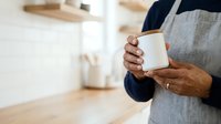 Mature man with medium brown skin tone wearing gray apron holding white ceramic mug in kitchen, suitable for coffee, beverage, hospitality, and lifestyle content.