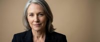 Mature woman with gray hair wearing dark blazer against neutral beige background, professional headshot for corporate, business, and editorial use.