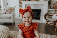 Smiling baby girl in rust-colored ruffle dress and matching bow headband sitting on floor, bright natural light from window, cozy living room with fireplace in background.