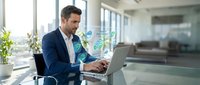 Young male professional with dark hair and facial stubble, wearing navy blazer and light shirt, working on laptop at modern office desk with green plant and window view, representing business technology and corporate work environment.