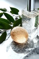 Elegant glass perfume bottle with golden bath bomb and green leaves on marble surface, creating luxurious spa and beauty composition for cosmetic advertising.