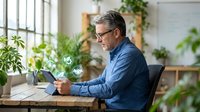 Mature Caucasian businessman using tablet with glowing green eco icons in plant-filled office, exploring sustainable technology solutions.