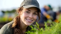 Young Caucasian woman in baseball cap smiling outdoors in natural setting with soft bokeh background, casual lifestyle portrait showing genuine happiness and vitality.