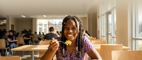 Young African American woman with dreadlocks enjoying healthy meal in modern cafeteria, smiling while eating salad during lunch break at university or workplace.