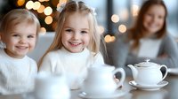 Young girls in white sweaters smiling at tea party with porcelain set and bokeh lights in background, mother watching from distance, cozy home atmosphere.