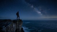 Man standing on rocky cliff edge overlooking ocean at night under starry sky with milky way galaxy visible, adventure and exploration concept for travel and nature themes.