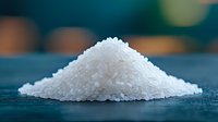 White granulated sugar pile on dark surface with blurred kitchen background. Raw ingredient for baking, cooking and food preparation in culinary applications.