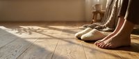 Cozy morning scene with bare feet on wooden floor next to knitted slippers in warm sunlight streaming through window creating peaceful home atmosphere.
