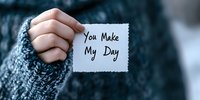Hand holding white note with handwritten message "You make my day" against blurred gray knit sweater background, creating intimate romantic moment.
