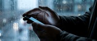 African American businessman hands holding smartphone with glowing screen in modern office environment during evening hours with city lights background.
