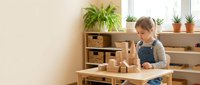 Young blonde girl playing with wooden building blocks at table in bright Montessori classroom with natural materials and plants creating educational learning environment.