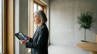 Professional mature Caucasian businesswoman using digital tablet in modern office interior with natural lighting and minimalist design elements.