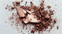 Crushed bronze eyeshadow powder with broken compact pieces scattered on light background, creating makeup texture for cosmetic product photography.
