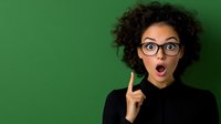 Young African American woman with curly hair and glasses showing surprised expression with raised finger against green background. Concept of idea or eureka moment.