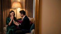 Young Caucasian man in black suit proposing to surprised woman in green dress with engagement ring in elegant hotel room interior.