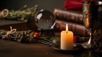 Mystical fortune telling setup with burning candle, crystal ball, ancient books and ornate chalice on wooden table creating atmospheric divination scene.