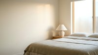 Serene bedroom interior with unmade bed, minimalist table lamp, and natural light streaming through window for home decor.