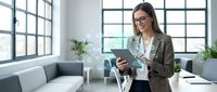 Young woman with dark hair and glasses, wearing a brown blazer and white blouse, holding a tablet while standing in a modern office space with gray sofa, potted plants, and large windows.