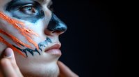 Side profile of young woman with creative blue and orange face paint makeup against dark background, artistic fashion portrait with dramatic lighting.