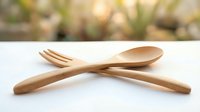 Wooden fork and spoon crossed on white surface with blurred green garden background, eco-friendly sustainable kitchen utensils for dining and cooking concepts.