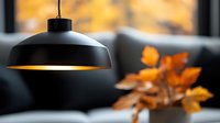 Modern black pendant lamp illuminating cozy living room interior with autumn leaves decoration, creating warm atmospheric lighting against blurred background.