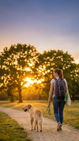 Young woman with blonde hair walking golden retriever on path at sunset, wearing backpack and casual clothing for outdoor recreation and pet care concepts.