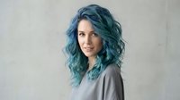 Young woman with vibrant blue-green wavy hair smiling confidently against neutral background for lifestyle and beauty concepts.
