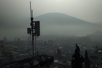 Moody cityscape with cell tower silhouette against misty mountain backdrop, dark atmospheric urban landscape in foggy weather conditions at dusk.
