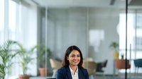 Young professional woman with dark hair and medium skin tone wearing navy blazer and white shirt, smiling in modern glass-walled office with plants and natural light, suitable for corporate business and workplace content.