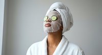 Woman in white bathrobe and towel enjoying spa facial mask with cucumber slices on eyes for skincare routine and relaxation at home wellness treatment.