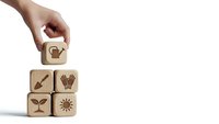 Hand placing wooden cube with watering can icon onto stack of blocks featuring gardening symbols on white background for eco-friendly design concepts.