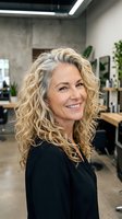 Confident mature blonde businesswoman smiling in modern office workspace with curly hair and professional attire for corporate and career success concepts.