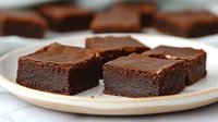 Rich, fudgy chocolate brownies with glossy ganache topping served on white plate. Dense, moist texture and dark chocolate appearance create indulgent dessert presentation.