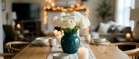 Elegant dining room table setting with white roses in teal vase, warm string lights creating cozy atmosphere for dinner party or special occasion celebration.