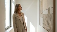 Elegant middle-aged Caucasian businesswoman in beige suit contemplating abstract art in a sunlit modern gallery space.