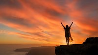 Woman celebrating success with raised arms on mountain cliff edge during dramatic orange sunset sky over ocean landscape for motivation concepts.