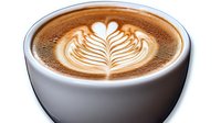 Creamy latte art in white ceramic cup showing detailed rosetta pattern with heart design on top of rich brown coffee foam, close-up view on white background.