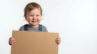 Happy toddler boy holding blank cardboard sign with big smile wearing striped shirt and overalls on white background for advertising mockup design.
