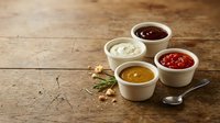 Four colorful condiment sauces in white bowls on rustic wooden table with herbs and spoon for culinary cooking and food preparation concepts.