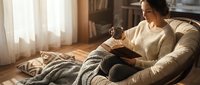 Young woman relaxing on couch reading book while drinking coffee in cozy living room with warm natural lighting creating peaceful morning atmosphere.