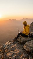 Woman in yellow jacket and beanie sitting on mountain cliff with laptop at sunrise, working remotely with valley landscape below. Remote work, freelance, digital nomad concept.